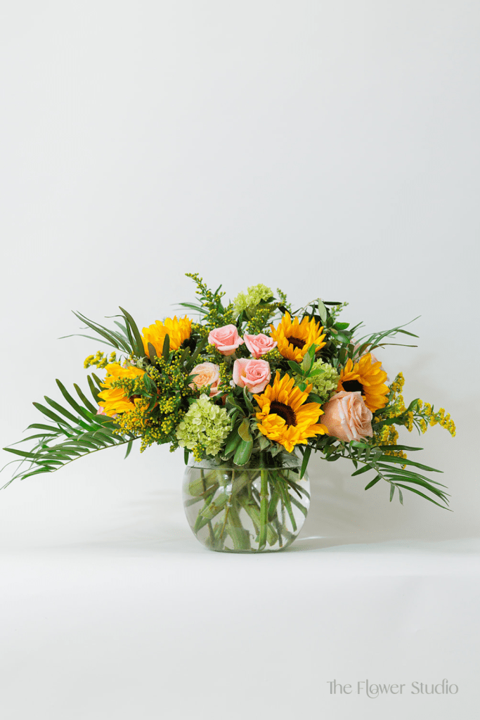 Radiant and full of warmth, this vibrant arrangement brings together the cheerful beauty of Sunflowers with the soft elegance of Spray Roses and Mini Hydrangeas.