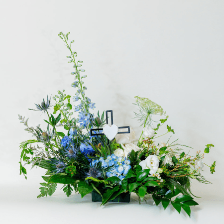 This striking sympathy arrangement combines soothing blues and crisp whites with a touch of purple, creating a sense of calm and reverence with a cross.