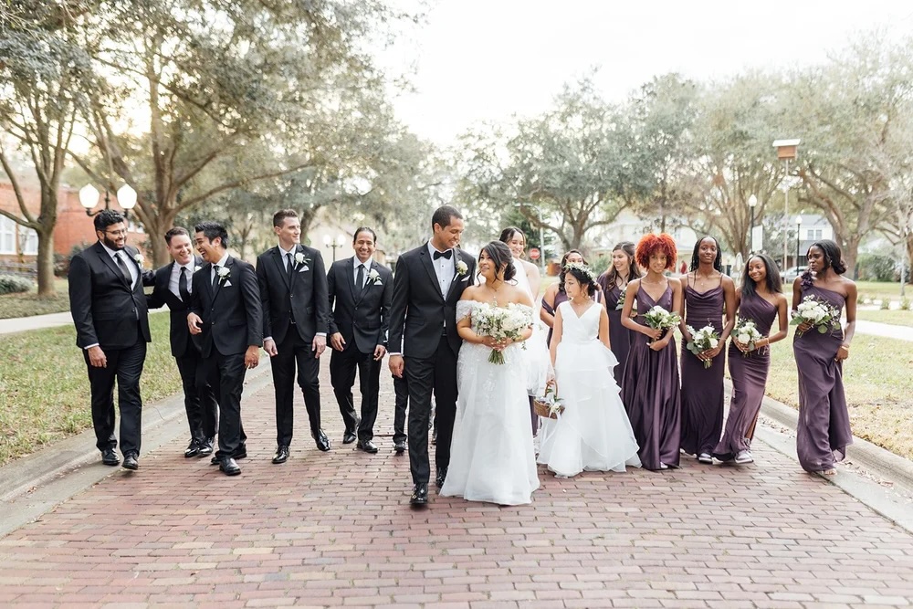 A joyful wedding party walking together down a brick pathway, with the bride in a white gown and bridesmaids in purple dresses.
