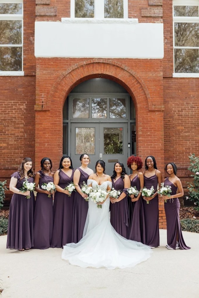 Bride and bridesmaids posing outside Venue 1902, wearing elegant purple dresses. The bride is in a white gown holding a bouquet of white flowers, while the bridesmaids are holding smaller bouquets.