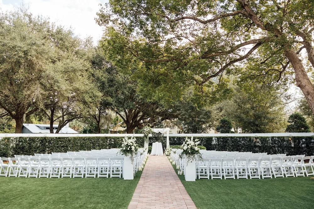 An outdoor wedding ceremony setup featuring white chairs arranged along a paved pathway leading to a floral-decorated altar, surrounded by lush greenery and large trees.