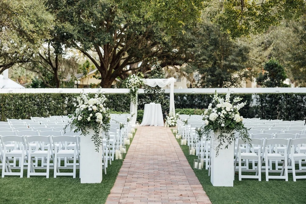 A beautifully arranged outdoor wedding ceremony setup featuring white chairs lined along a brick path, adorned with white floral arrangements on pedestals, and a decorated altar under a canopy of greenery and trees.