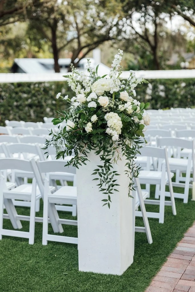 A tall floral arrangement featuring white flowers and lush greenery stands next to rows of white chairs set up for a wedding ceremony, on a neatly trimmed lawn.