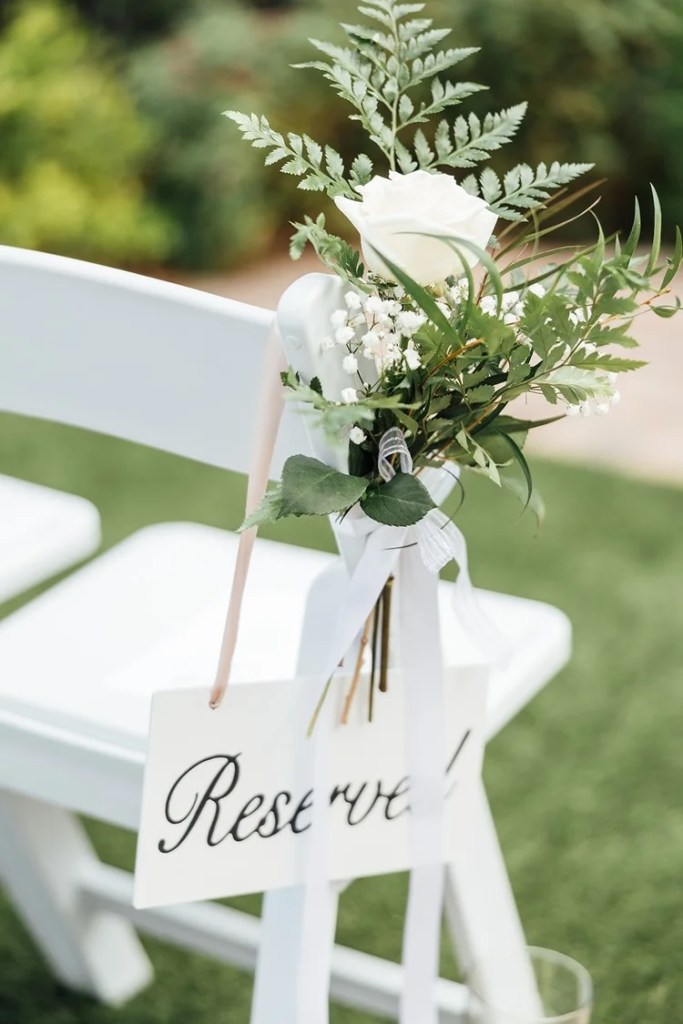 A close-up of a wedding ceremony chair decorated with a white rose bouquet, greenery, and a sign that says 'Reserved'.