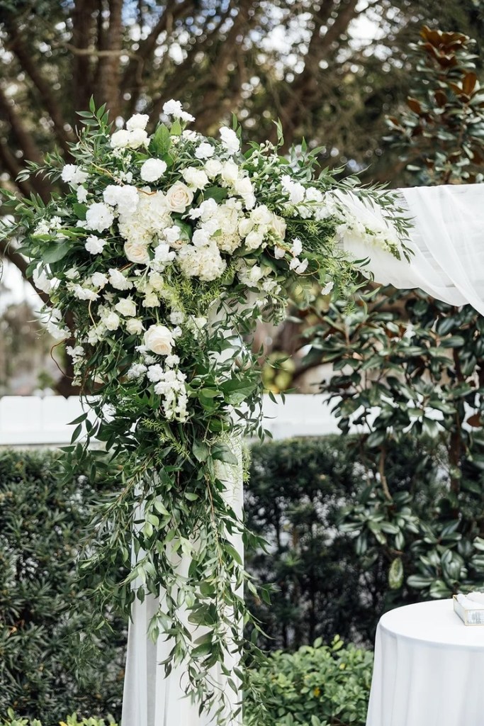 An elegant wedding ceremony arch adorned with white flowers and lush greenery, set against a backdrop of trees.
