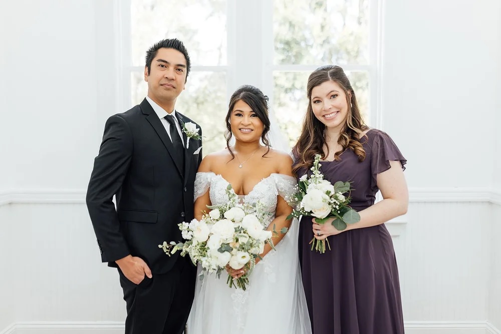 A bride in a beautiful wedding dress poses with her groom and bridesmaid in a light-filled indoor setting.