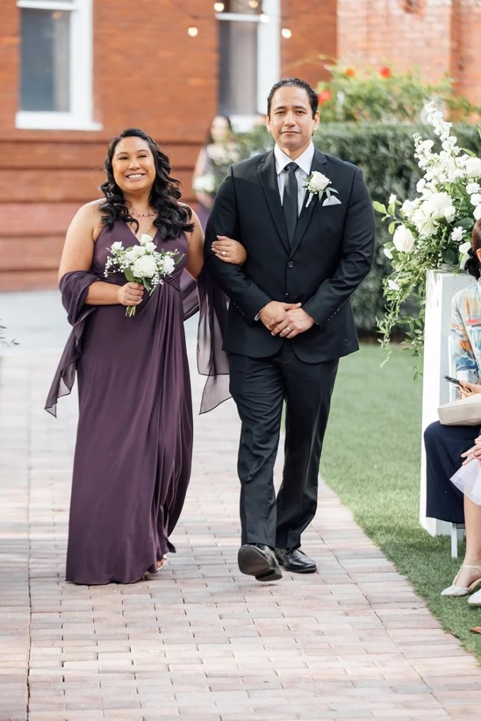 A woman in a purple dress holds a bouquet as she walks arm in arm with a man in a black suit during a wedding ceremony.
