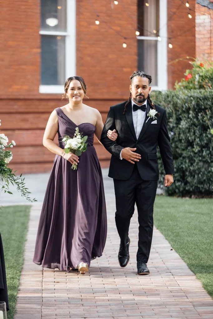 A woman in a purple gown and a man in a black tuxedo walk down a brick path together during a wedding ceremony, with greenery and string lights in the background.