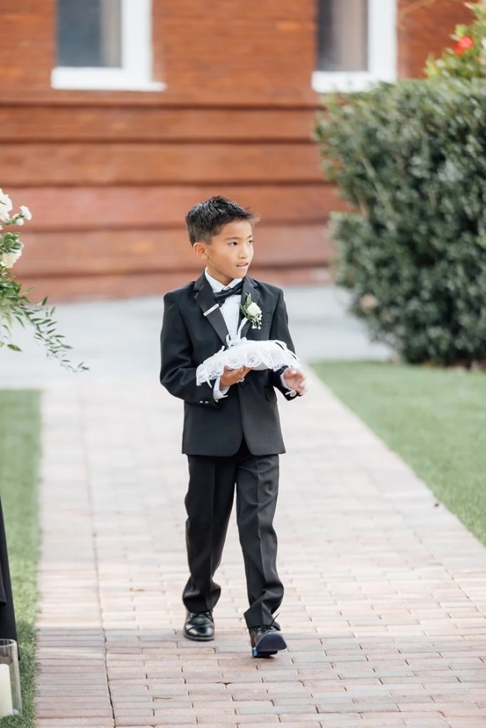 A young boy in a tuxedo with a bow tie walks down a brick path at a wedding, holding a decorative pillow.