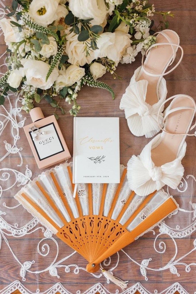 Wedding flat lay featuring a bouquet of white roses and greenery, a bottle of Gucci Bloom perfume, a vow book, and stylish sandals on a lace-patterned backdrop.