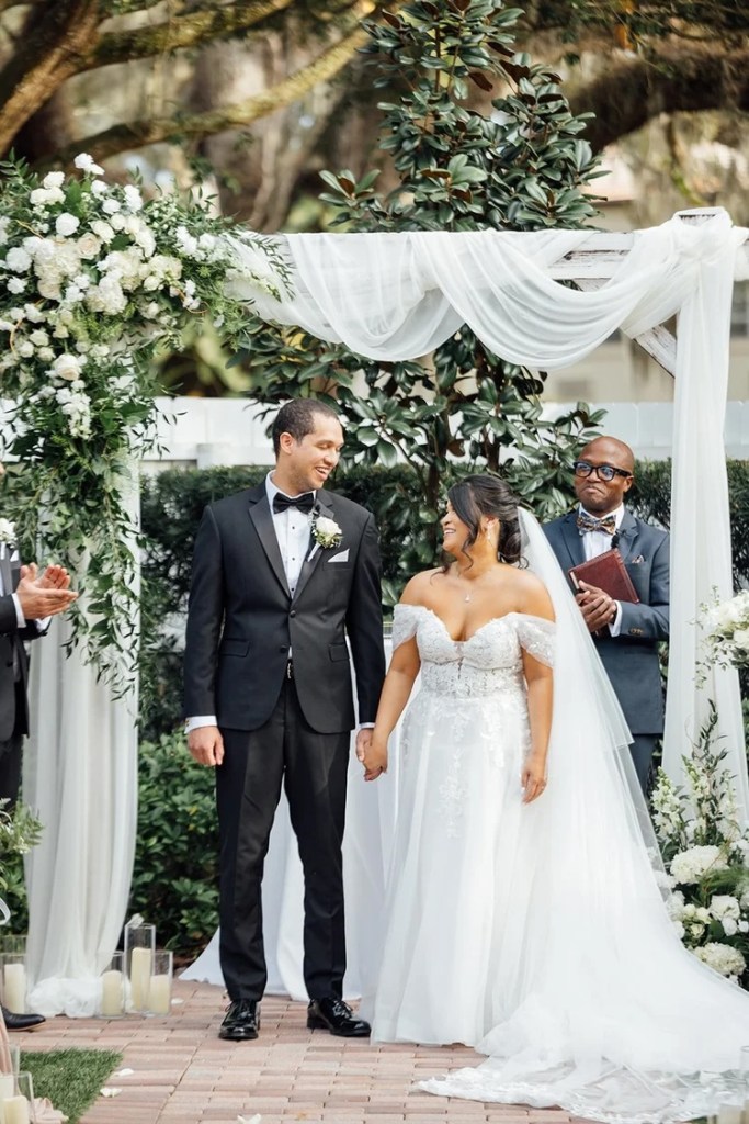 A joyful couple standing together during their wedding ceremony under a floral arch, with white flowers and greenery in the backdrop.