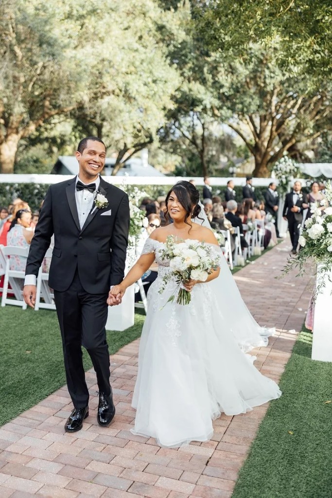 A happily married couple walks hand in hand down a picturesque outdoor aisle, surrounded by guests. The bride, in an elegant white gown holding a bouquet of white flowers, smiles at the groom, who is dressed in a classic black tuxedo. Lush greenery and white florals decorate the venue, creating a romantic and sophisticated atmosphere.