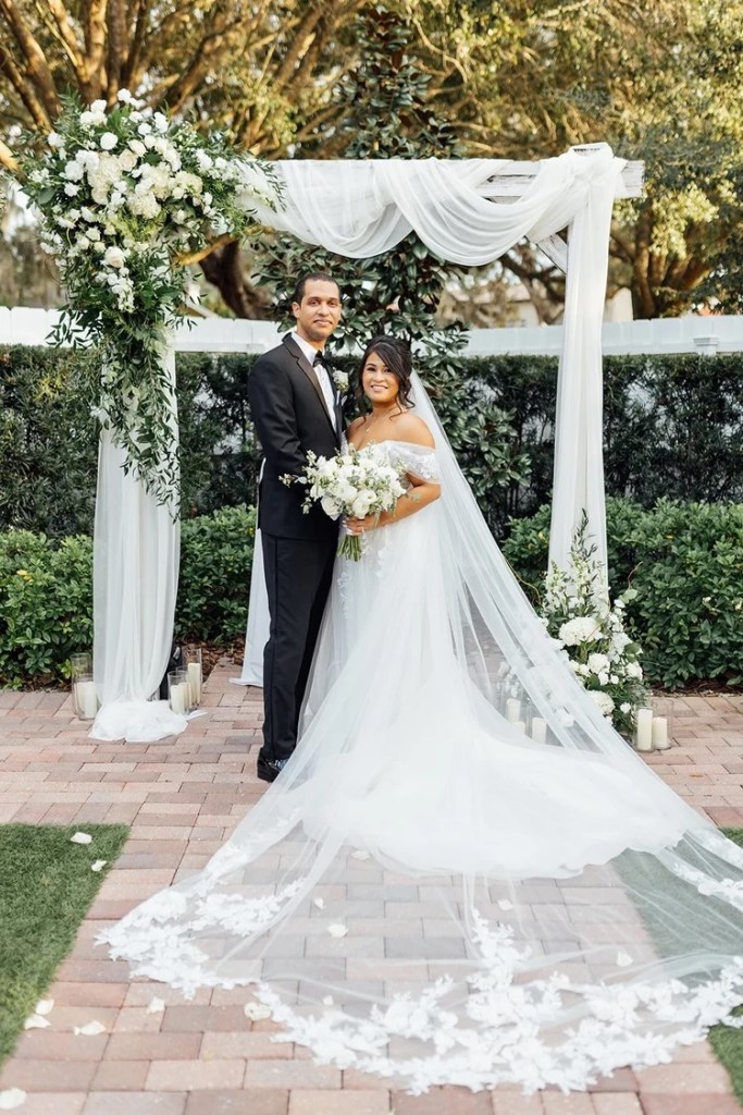 A couple stands together at an outdoor wedding ceremony, surrounded by greenery and white floral arrangements. The bride wears an off-the-shoulder wedding gown with a long, flowing veil, while the groom is in a formal black suit. An elegant floral arch decorates the background.