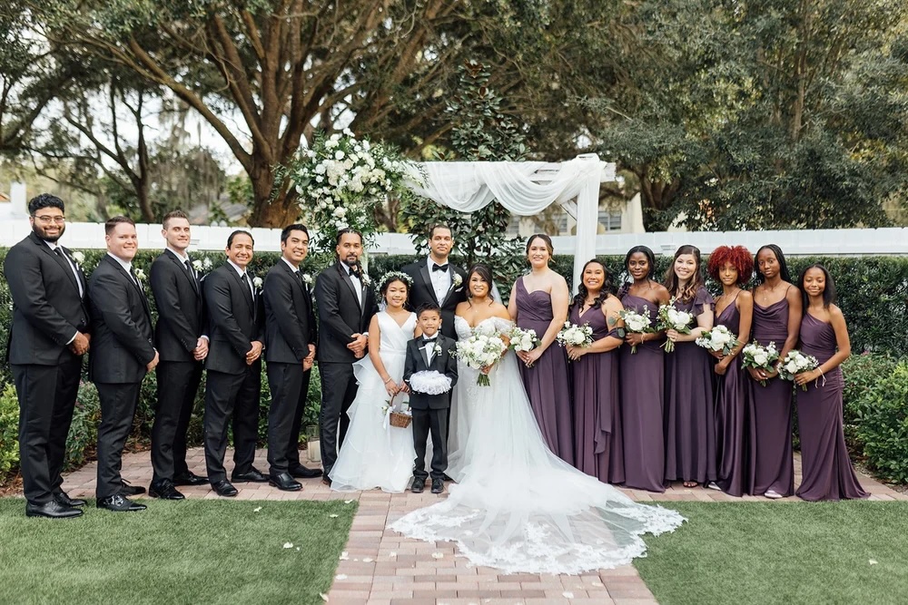 A wedding party poses together outdoors, featuring the bride in a white gown and veil alongside bridesmaids in purple dresses, and groomsmen in black suits. The backdrop includes floral decorations and greenery.