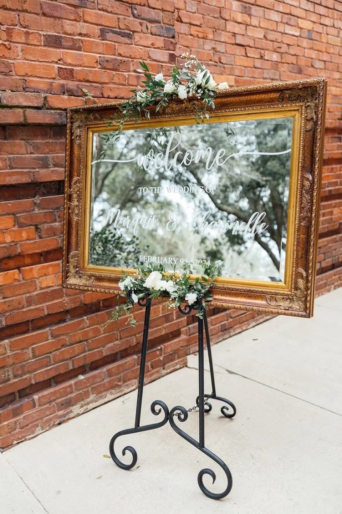A decorative welcome sign for a wedding, framed in an ornate gold frame with floral accents, displayed against a rustic brick wall.