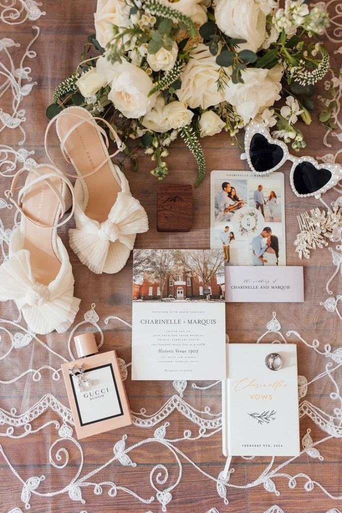 A flat lay arrangement featuring wedding essentials: elegant white roses bouquet, a pair of stylish sandals, a bottle of Gucci Bloom perfume, wedding invitation, vow book, and heart-shaped sunglasses on a lace background.
