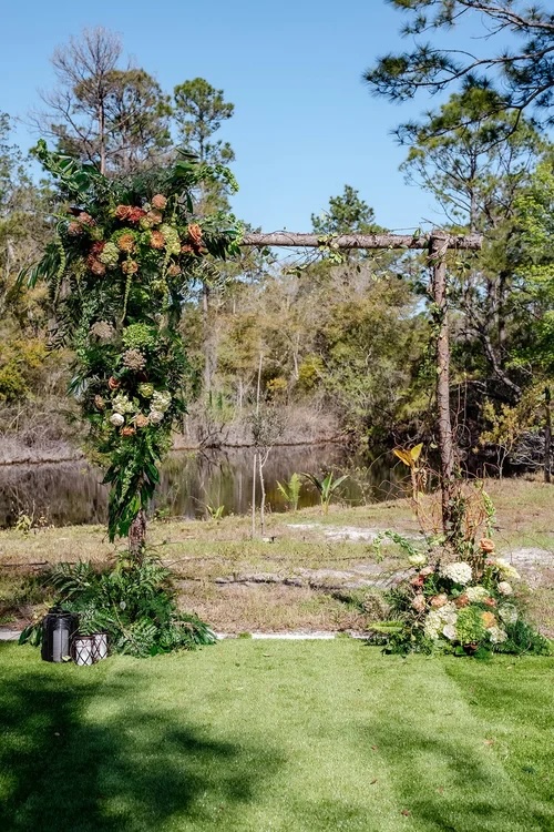 An elegant wedding arch adorned with lush greenery and vibrant floral arrangements, set in a picturesque outdoor location by a body of water.