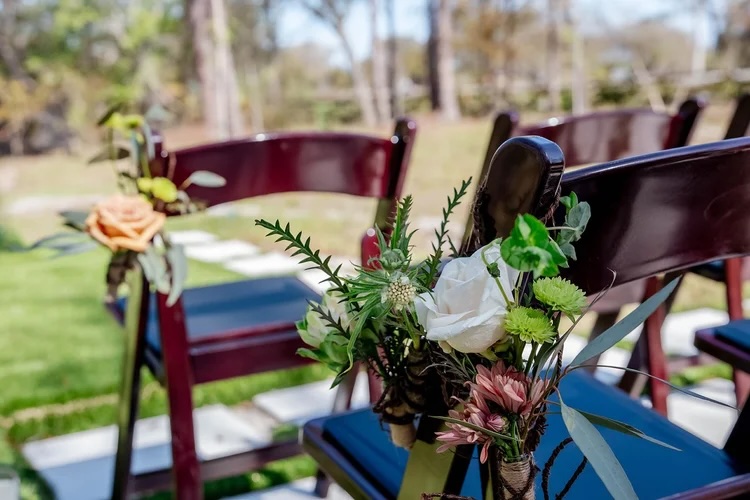Close-up of wedding ceremony chairs adorned with floral arrangements, including roses and greenery, set in a natural outdoor setting.