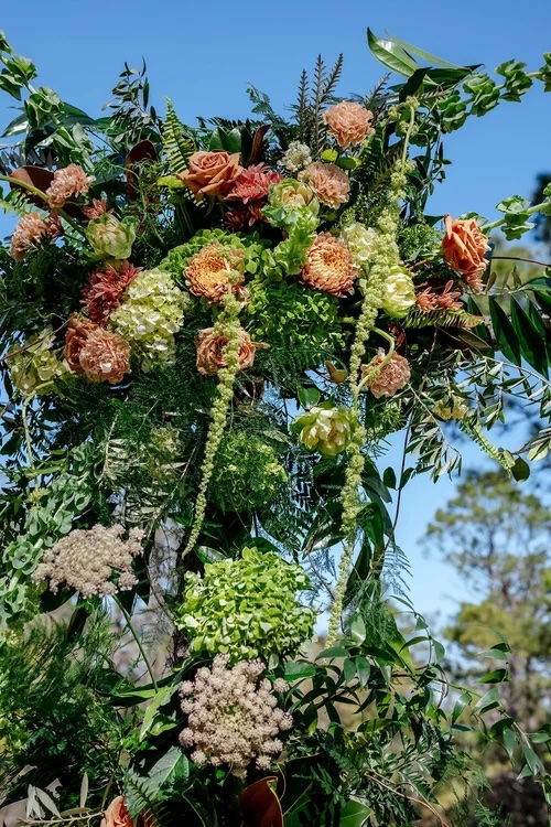 A beautiful floral arch featuring an array of lush blooms in shades of peach, green, and cream, adorned with various greenery against a clear blue sky.