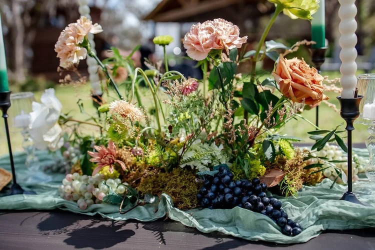 A beautifully arranged centerpiece featuring a variety of colorful flowers, including peonies and roses, paired with green foliage and decorative elements, set on a textured fabric backdrop.