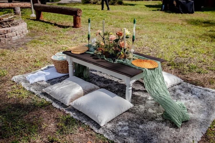 A beautifully styled outdoor table arrangement featuring a rustic wooden table adorned with a floral centerpiece, candles, and vibrant textiles, set on a grassy area with cozy floor cushions.