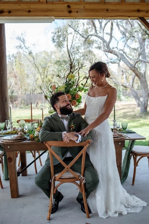 A bride and groom share a joyful moment at a beautifully styled wedding table surrounded by lush floral dรฉcor in a natural setting.