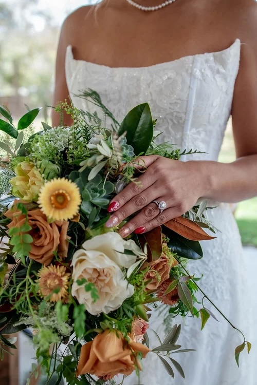 A close-up of a bridal bouquet featuring lush blooms in warm hues, held by a bride wearing a strapless wedding dress, showcasing her engagement ring.