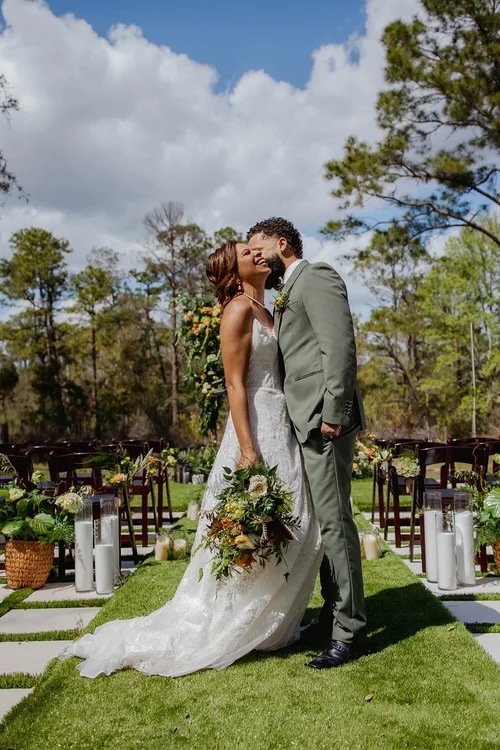 A couple kisses at their wedding ceremony, surrounded by lush greenery, floral arrangements, and elegant decor under a partly cloudy sky.
