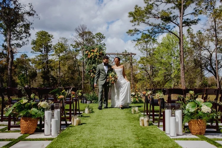 A couple walks down a lush green aisle towards an outdoor wedding altar surrounded by trees and floral decorations.