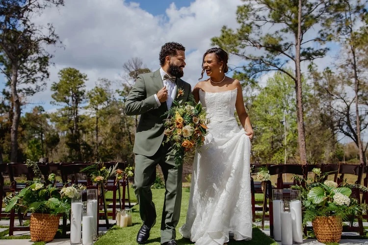 A groom and bride walk hand in hand down an outdoor aisle, surrounded by greenery and floral arrangements. The bride holds a bouquet of colorful flowers and wears a white wedding dress, while the groom is dressed in a green suit. Soft, cloudy skies and lush trees create a romantic backdrop.