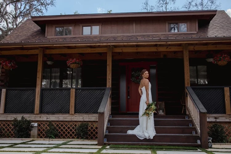 A bride in a white wedding dress stands on the steps of a charming wooden cabin, holding a lush bouquet of flowers, smiling back at the camera. The setting features natural elements and floral decorations, enhancing the romantic atmosphere.