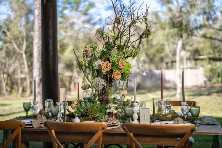 A beautifully styled table centerpiece featuring a tall floral arrangement with roses, greenery, and hanging glass ornaments, set outdoors with an elegant table setting and natural surroundings.