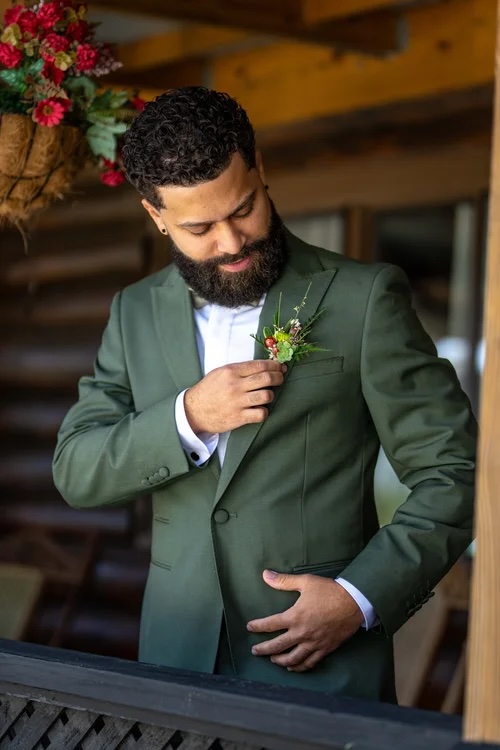 A man in a green suit adjusts a boutonniere on his jacket while standing in a rustic setting. Lush blooms can be seen in the background, enhancing the romantic atmosphere.