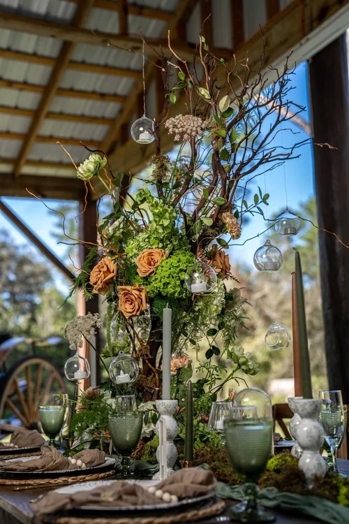 A beautifully styled table arrangement featuring lush floral designs with blooming roses, greenery, and hanging glass orbs, set in a rustic outdoor venue.