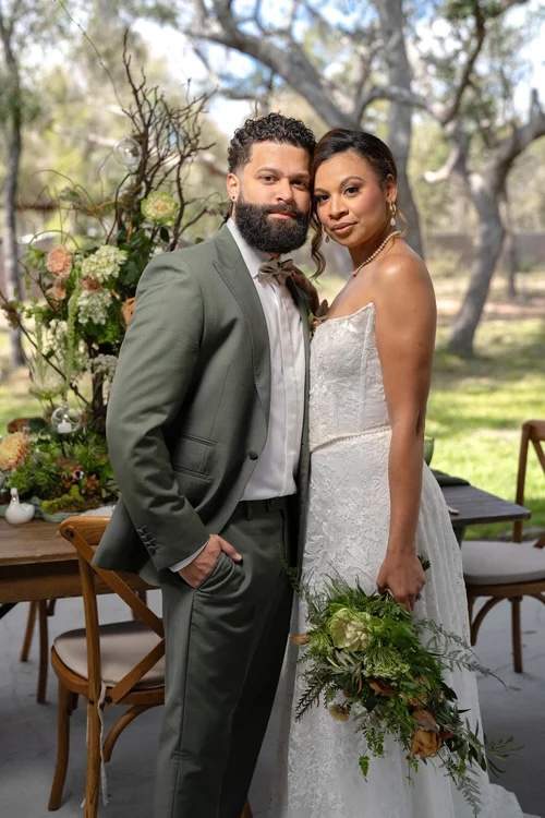 A couple stands together in a natural setting, surrounded by lush greenery and floral arrangements, showcasing a rustic wedding theme.
