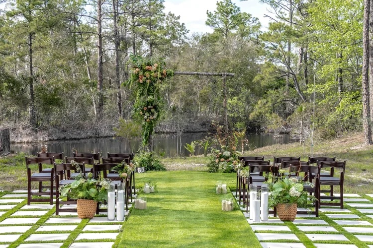 Outdoor wedding ceremony setup featuring wooden chairs arranged along a green aisle with a floral arch at the end, surrounded by nature and a small body of water.