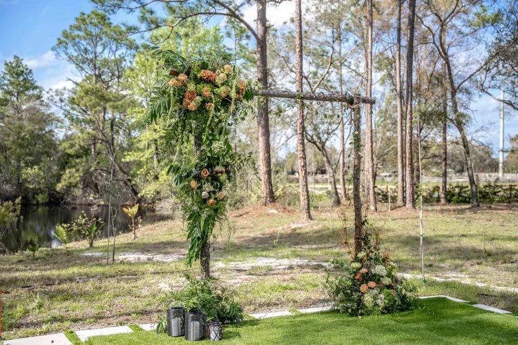 A beautifully designed outdoor wedding arch adorned with lush greenery and soft peach-colored flowers, set in a natural landscape near a body of water.