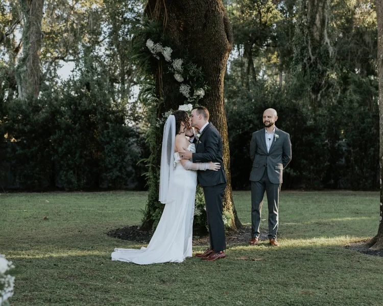 A radiant bride in an elegant white gown kisses her groom under a beautifully adorned tree, surrounded by lush greenery, while a man in a suit stands in the background, capturing the heartfelt moment of their wedding ceremony.