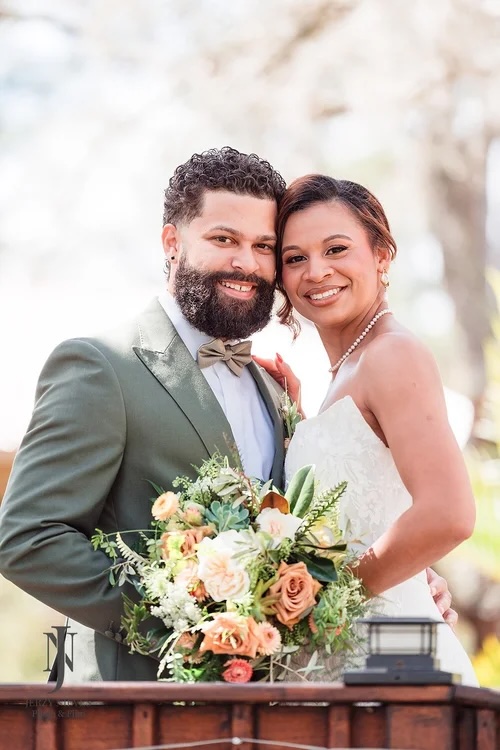 A couple joyfully posing together at an outdoor wedding, surrounded by natural beauty. The bride holds a bouquet filled with soft-colored blooms, while the groom sports a stylish green suit and a bow tie.