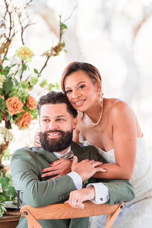 A couple posed together in a romantic setting, displaying affection and joy. The man has a beard and is dressed elegantly in a green suit, while the woman wears a beautiful wedding dress. They are surrounded by lush floral arrangements, enhancing the garden-themed ambiance.