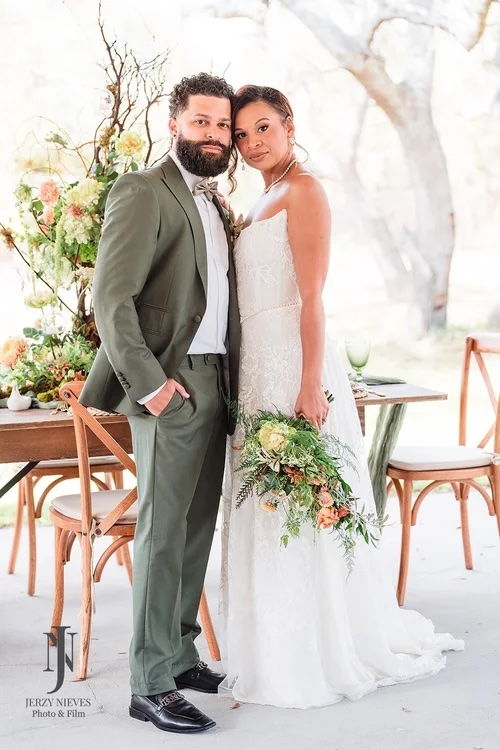 A bride and groom stand together in a garden setting, with the bride holding a bouquet of flowers and wearing a white lace dress, and the groom dressed in a green suit. Their expressions convey love and elegance, surrounded by floral arrangements in the background.