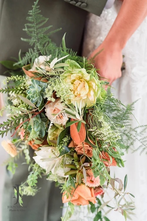 A close-up image of a bridal bouquet featuring a variety of lush blooms and greenery, including peonies, succulents, and ferns, held by a person wearing a wedding dress.