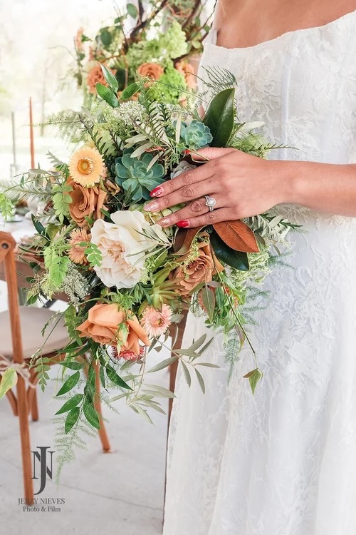 A close-up of a bride holding a lush bouquet featuring a mix of orange flowers, white blooms, green succulents, and greenery, with a delicate wedding dress in the background.