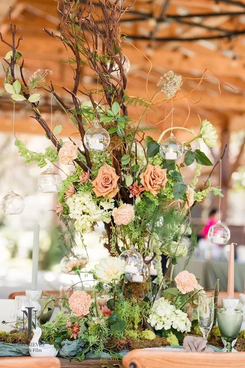 A beautifully arranged floral centerpiece featuring peach roses, white hydrangeas, and greenery, with hanging glass orbs, resting on a rustic table setting in a natural venue.