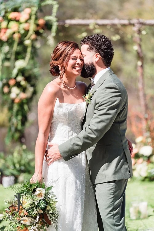 A joyful couple sharing a tender moment during their outdoor wedding ceremony, surrounded by lush greenery and floral arrangements.