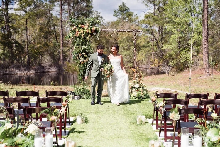 A happy couple walking down the aisle in a beautiful outdoor wedding setup, surrounded by lush greenery and floral decorations.