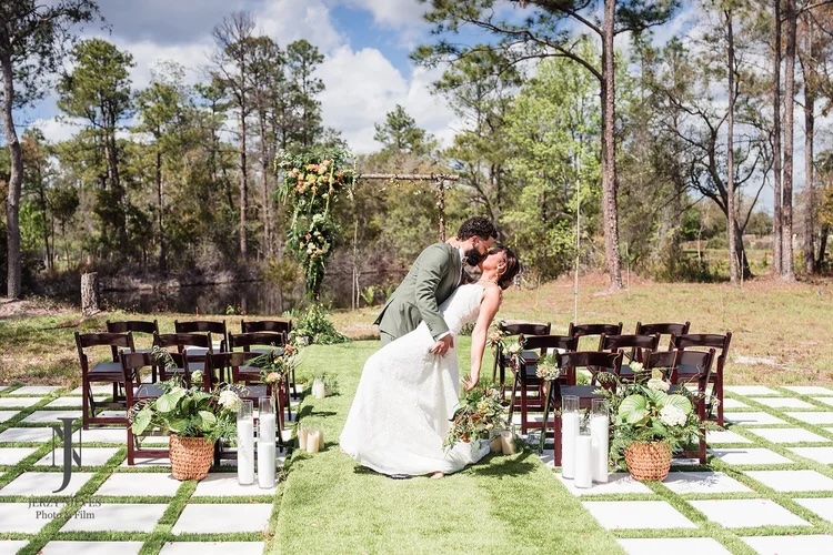 A couple shares a romantic kiss during their outdoor wedding ceremony, surrounded by lush greenery and elegantly arranged chairs. The scene features a decorative arch adorned with greenery and flowers, creating a picturesque setting.