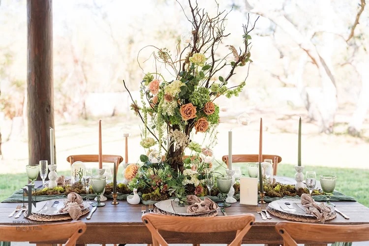 A beautifully styled outdoor table arrangement featuring lush floral centerpieces, elegant table settings with plates and glasses, and soft textured linens, set in a charming natural environment.