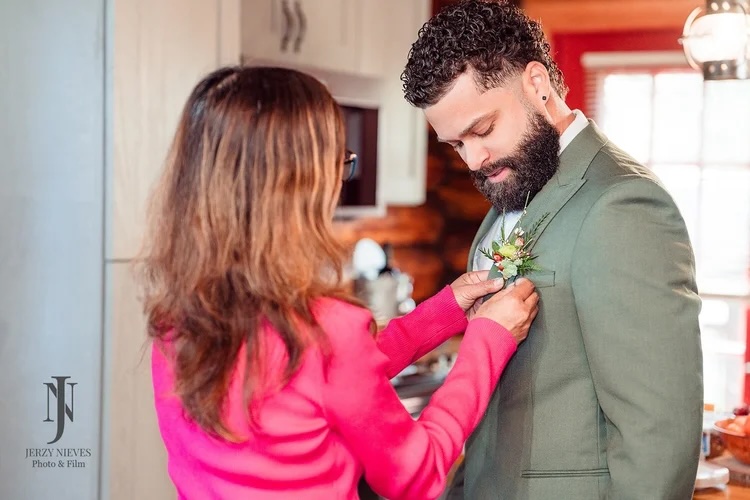 A woman carefully pins a floral boutonniere to the lapel of a man in a green suit, set in a cozy, rustic kitchen.