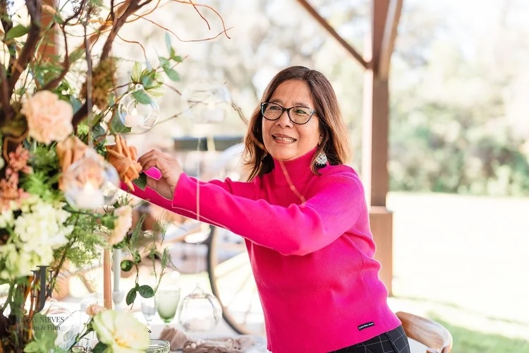 A smiling woman in a bright pink sweater arranging flowers and decorations at an outdoor event setup.